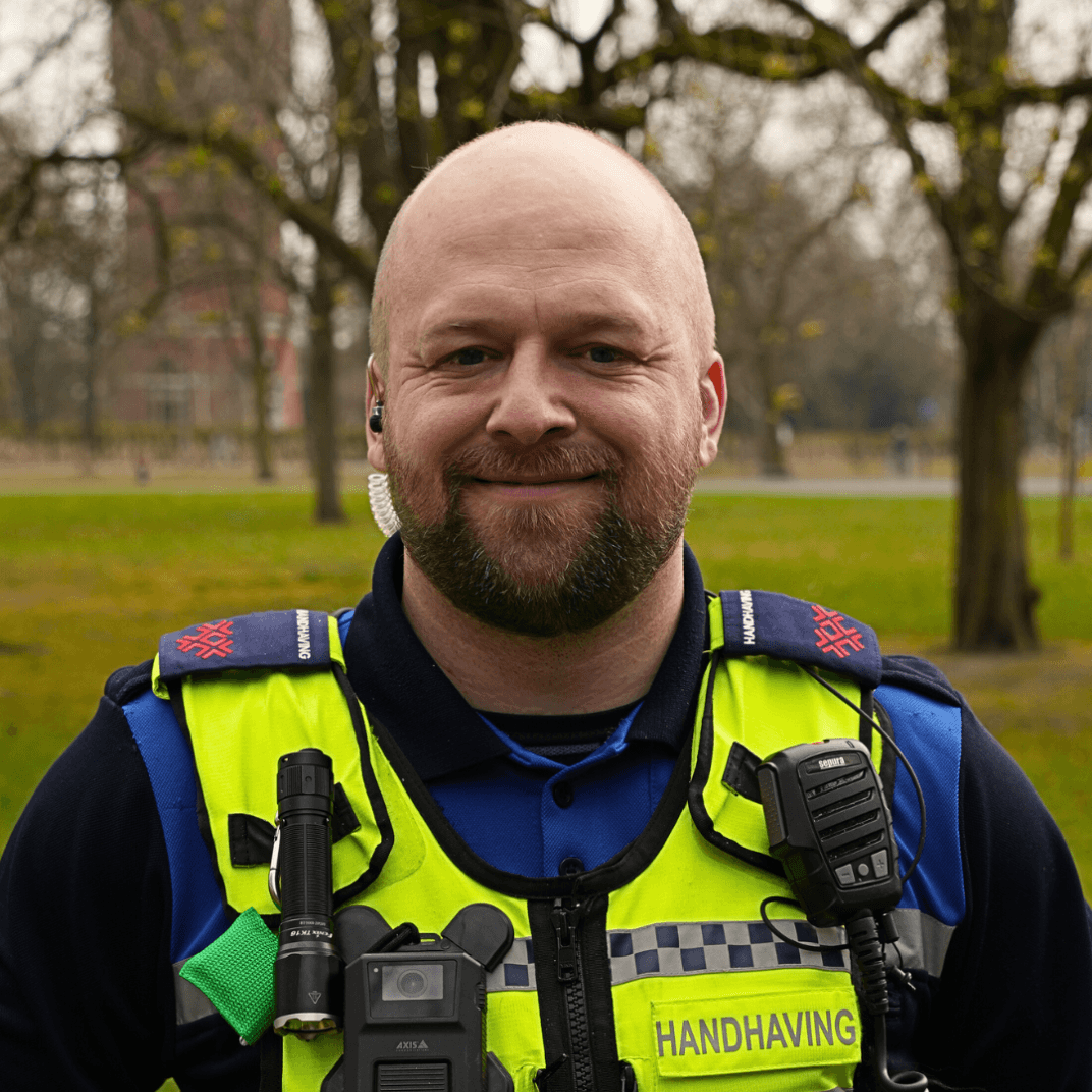 Wijkboa Michael in uniform in het park, zichtbare veiligheidsuitrusting, lachend in de camera.