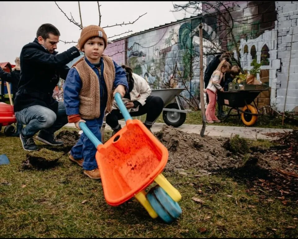 Kinderen en ouders planten bomen in de wijk; een jongen duwt een speelgoedkruiwagen tijdens een buurtvergroeningsactie.